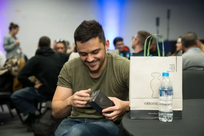 Homem sorrindo desembrulhando um presente em uma sala com várias pessoas ao fundo